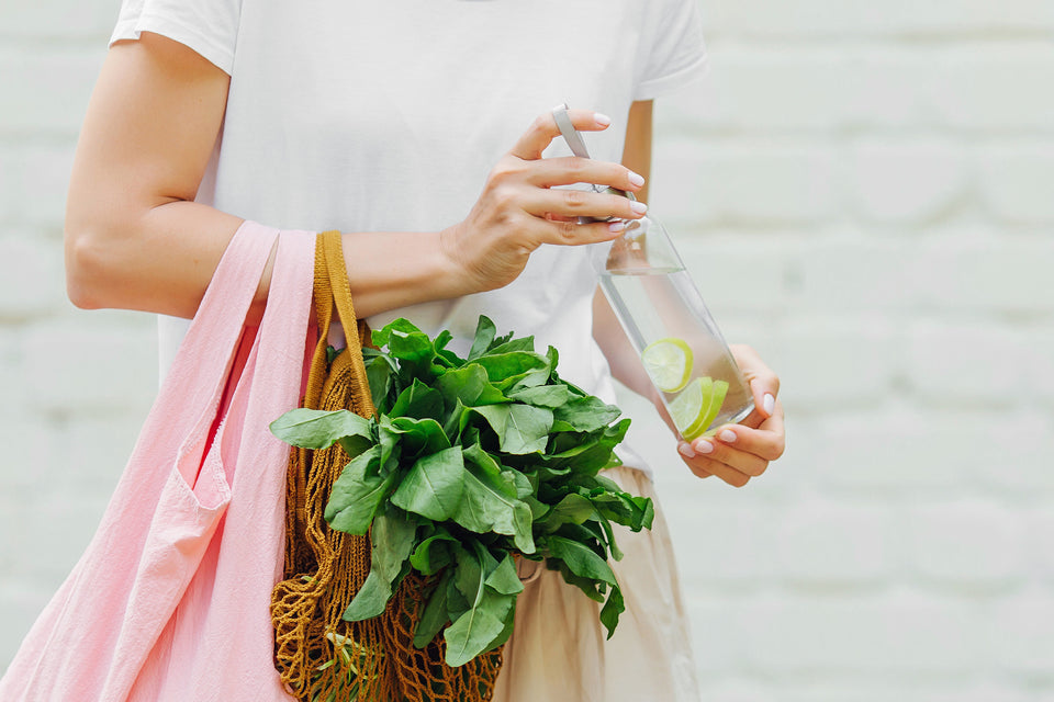 Someone carrying sustainable grocery bags filled with lettuce and in their hands is a glass water bottle and a reusable straw.