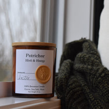 Load image into Gallery viewer, Next to an open window on a rainy day, our apothecary beeswax candle sits on the window ledge. A green and black sweater and linen pillow perch next to the candle
