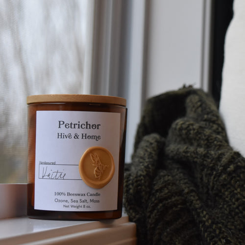 Next to an open window on a rainy day, our apothecary beeswax candle sits on the window ledge. A green and black sweater and linen pillow perch next to the candle