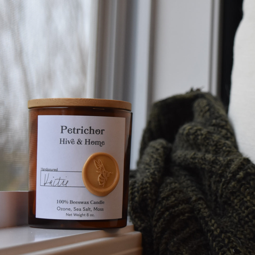 Next to an open window on a rainy day, our apothecary beeswax candle sits on the window ledge. A green and black sweater and linen pillow perch next to the candle