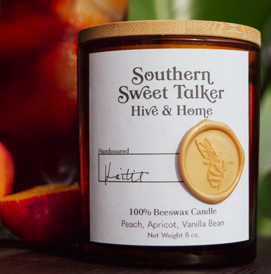 The Southern Sweet Talker candle in front of green foliage, an iced sweet peach tea, and a halved Southern peach.