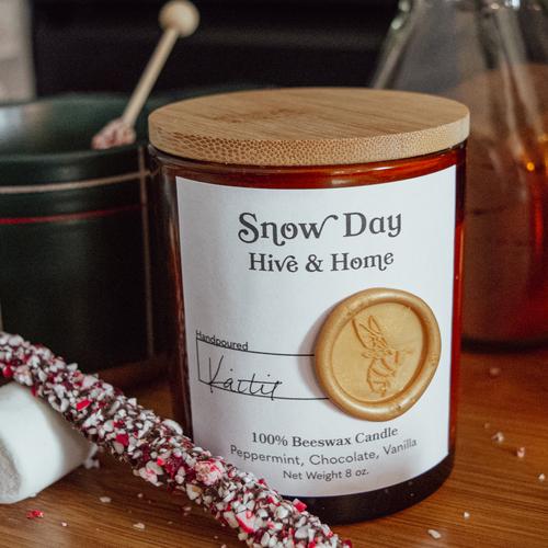 Foreground: Snow Day amber glass candle jar, a chocolate peppermint stir stick resting on a marshmallow. Background: left, dark green mug with red and while plaid stripes on it. Another stir stick is in the mug of hot cocoa. Right, jar of cocoa mix. Behind all of that is a fireplace.