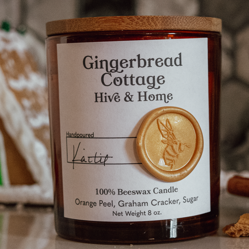 Foreground: Gingerbread Cottage amber glass candle jar. Background: gingerbread A-frame house, cinnamon sticks, and a piece of broken gingerbread.